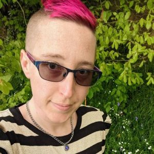 Picture of Alexis Bushnell, a white smiling lady with short pink hair, sunglasses, and a striped black-and-white shirt